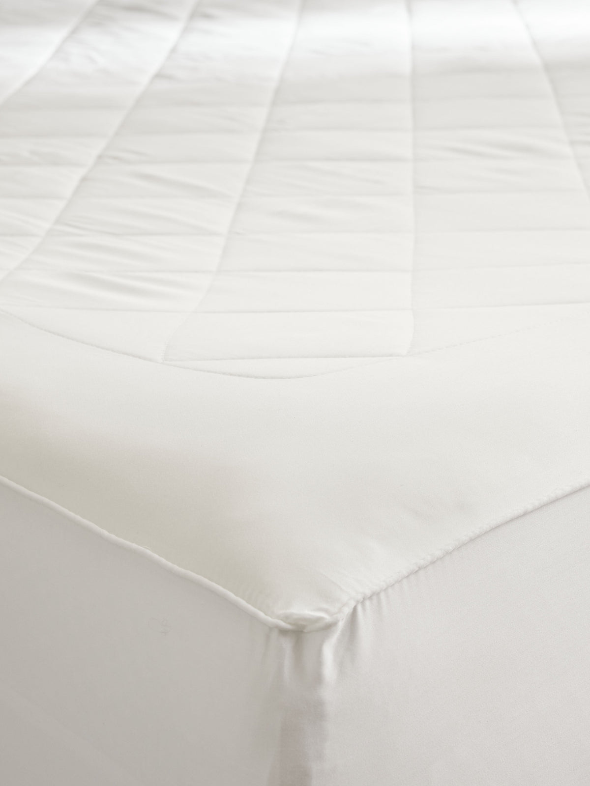 Detail of the mattress pad fitted around a mattress corner, showing the quilted top, smooth border, and fabric skirt wrapping down the sides.
