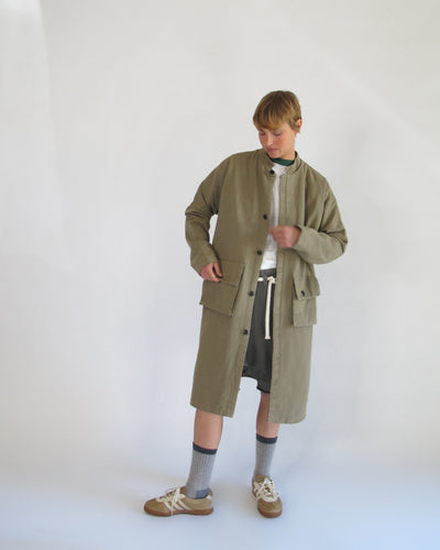 Model wearing an olive knee-length Passage Jacket over layered neutral clothing with gray socks and brown sneakers.
