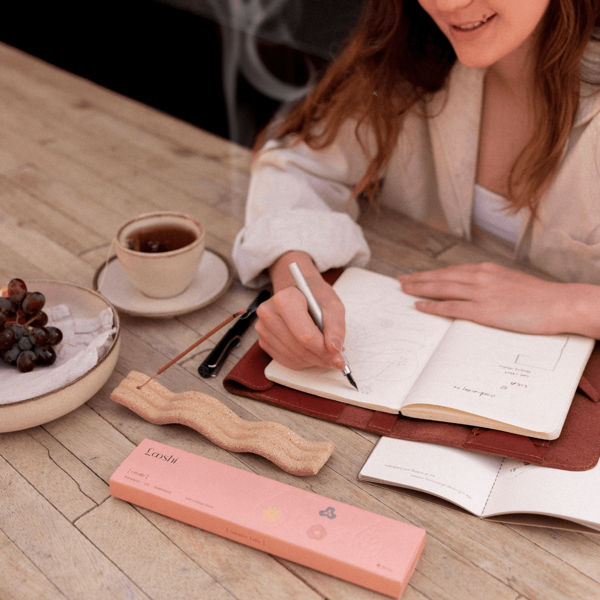 Woman writing in an open notebook at a wooden table with coffee and a peach box of Looshi Create incense sticks nearby