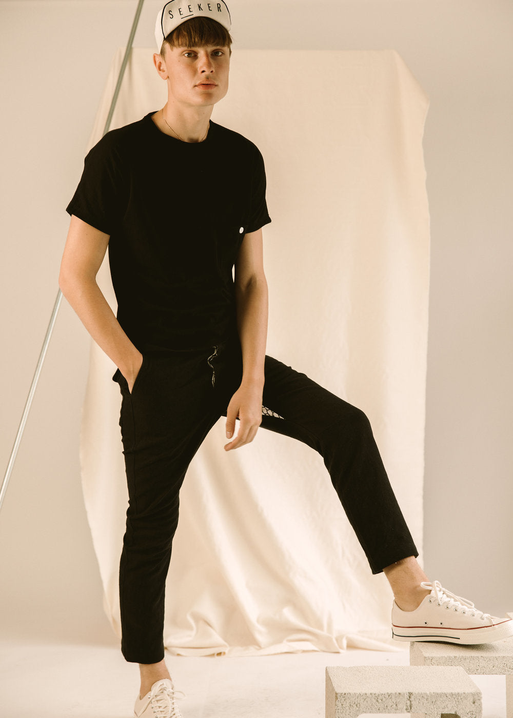 Model posing in a black Pocket Dolman Tee, slim black pants, white sneakers, and a baseball cap.