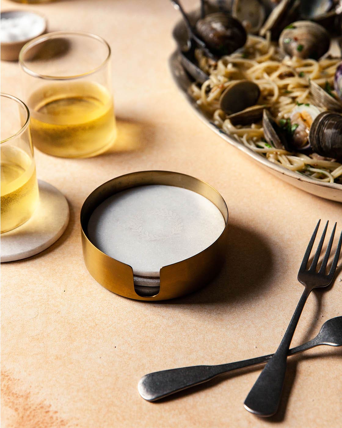 Brass holder of white marble coasters arranged on a dining table beside golden drinks, dark flatware, and a platter of food.