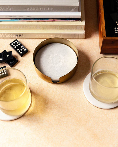 Overhead view of the marble coaster set among cocktail glasses, black dominoes, and a stack of books on a warm neutral tabletop.