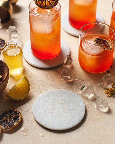 Loose white marble laurel coaster surrounded by colorful red-orange cocktails, citrus garnishes, ice cubes, and small bottles.