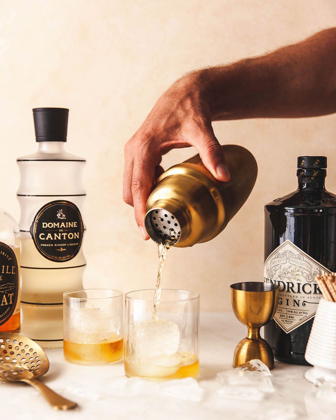 Hand pouring a pale cocktail from the brushed-gold shaker into two low glasses beside the matching jigger and liquor bottles.
