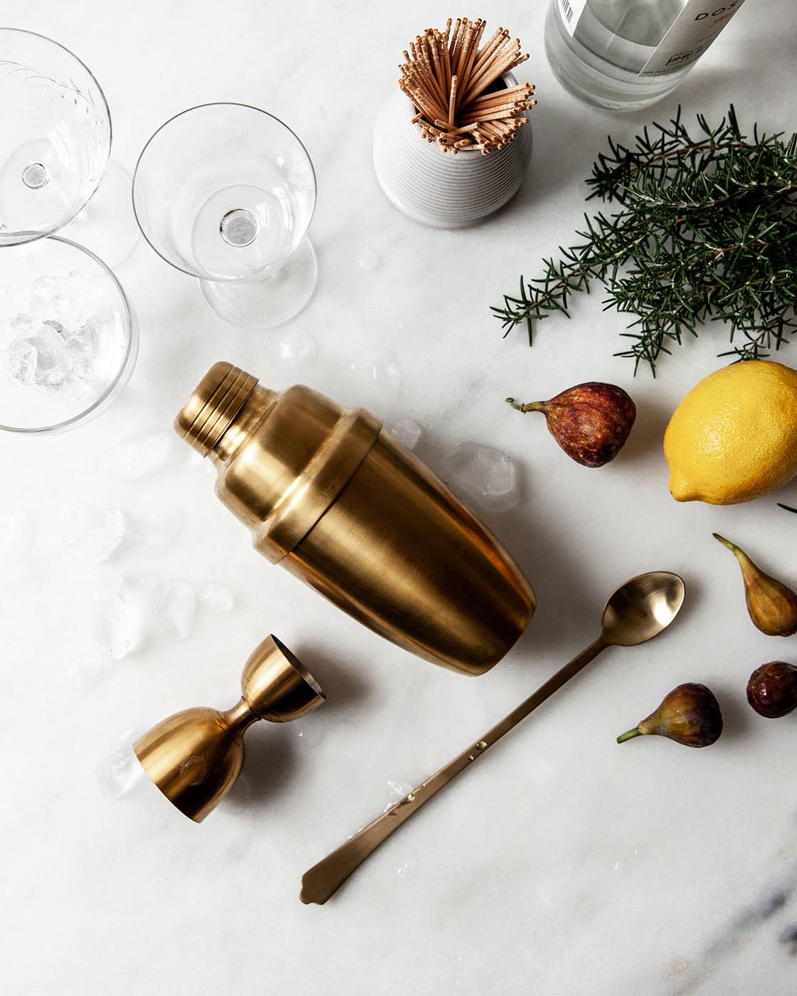 Overhead view of the gold cocktail shaker, jigger, strainer, and bar spoon styled with glassware, lemon, herbs, and scattered nuts.