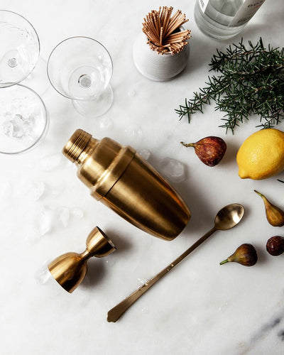 Overhead view of the gold cocktail shaker, jigger, strainer, and bar spoon styled with glassware, lemon, herbs, and scattered nuts.