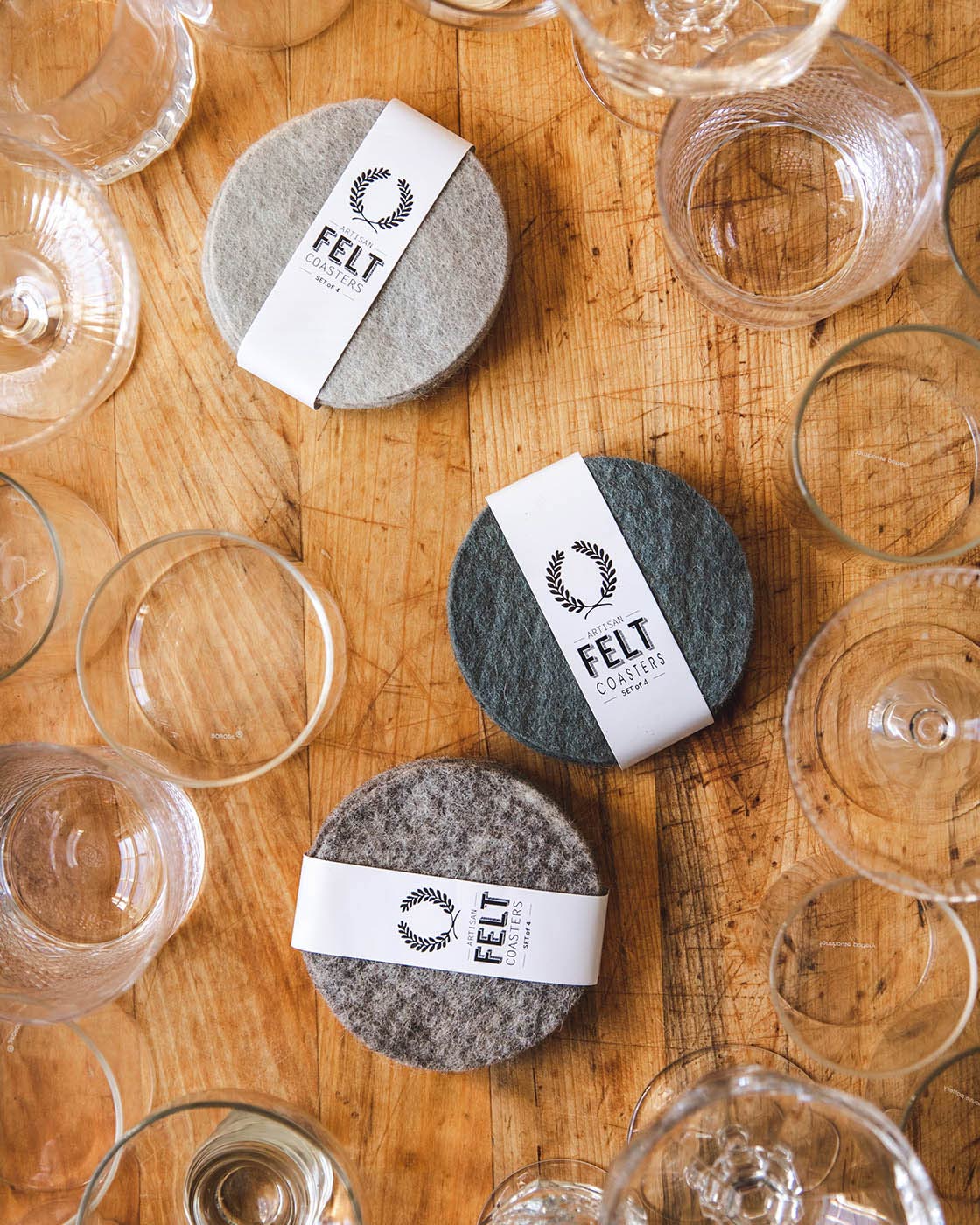 Overhead view of packaged gray felt coaster sets scattered among clear drinking glasses on a honey-toned wooden table.