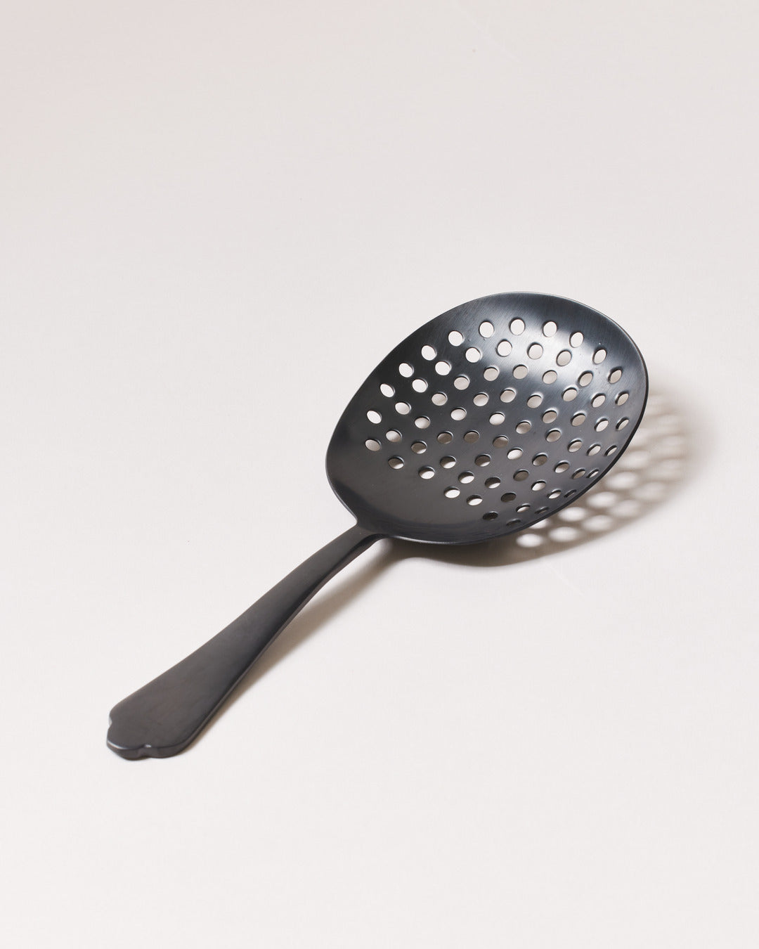 Brushed-onyx perforated cocktail strainer with a broad oval bowl, round drainage holes, and short decorative handle.