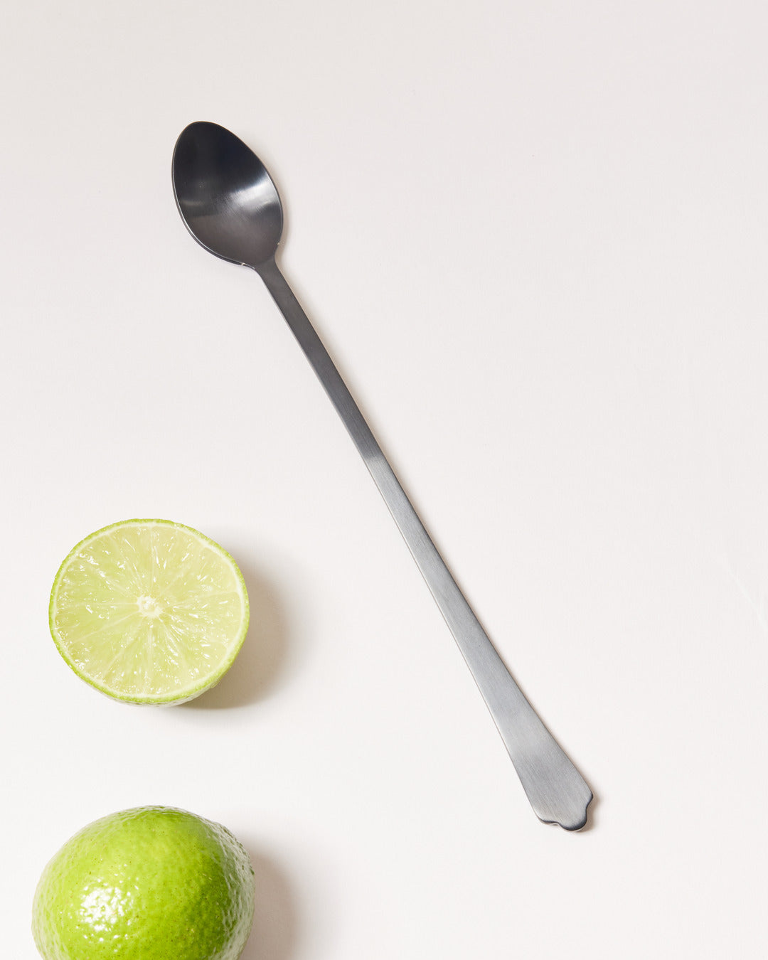 Long brushed-onyx bar spoon displayed diagonally beside a halved lime and a whole lime.