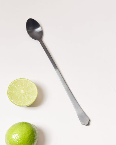 Long brushed-onyx bar spoon displayed diagonally beside a halved lime and a whole lime.