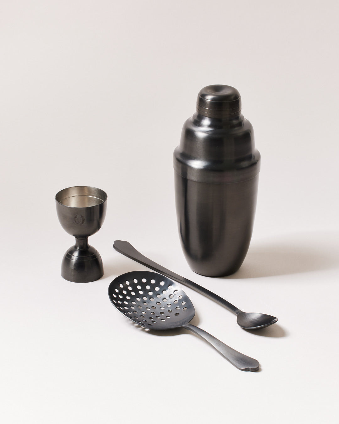 Four-piece brushed-onyx barware set with a dark charcoal cocktail shaker, double-sided jigger, perforated cocktail strainer, and long-handled bar spoon.