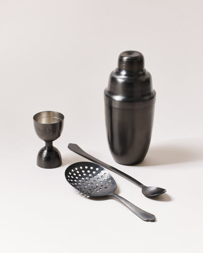 Four-piece brushed-onyx barware set with a dark charcoal cocktail shaker, double-sided jigger, perforated cocktail strainer, and long-handled bar spoon.