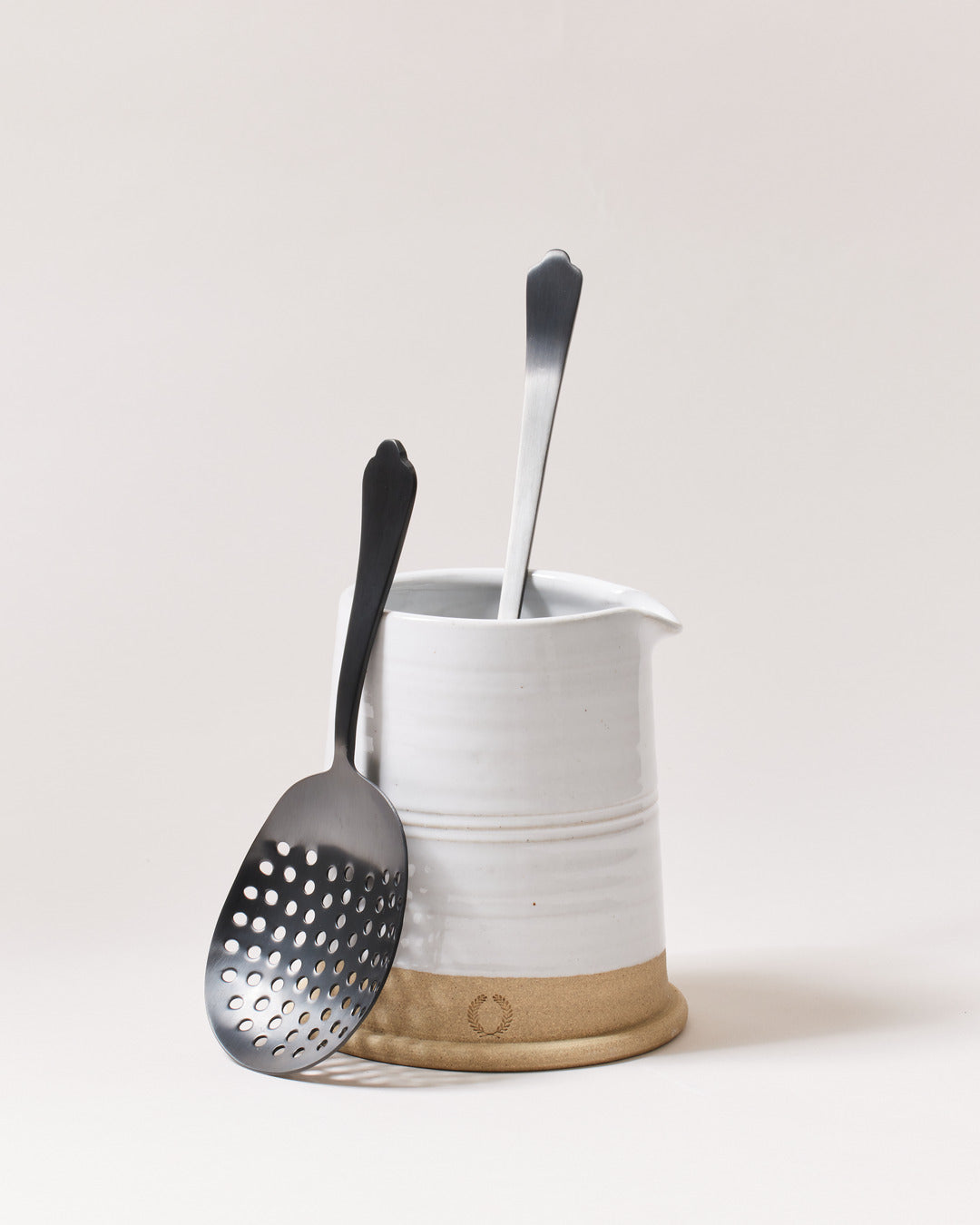 Onyx strainer and bar spoon resting in and against a tall white ceramic mixing vessel.