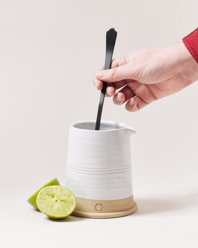 Hand stirring inside a white ceramic mixing vessel with the long onyx bar spoon, with a halved lime nearby.
