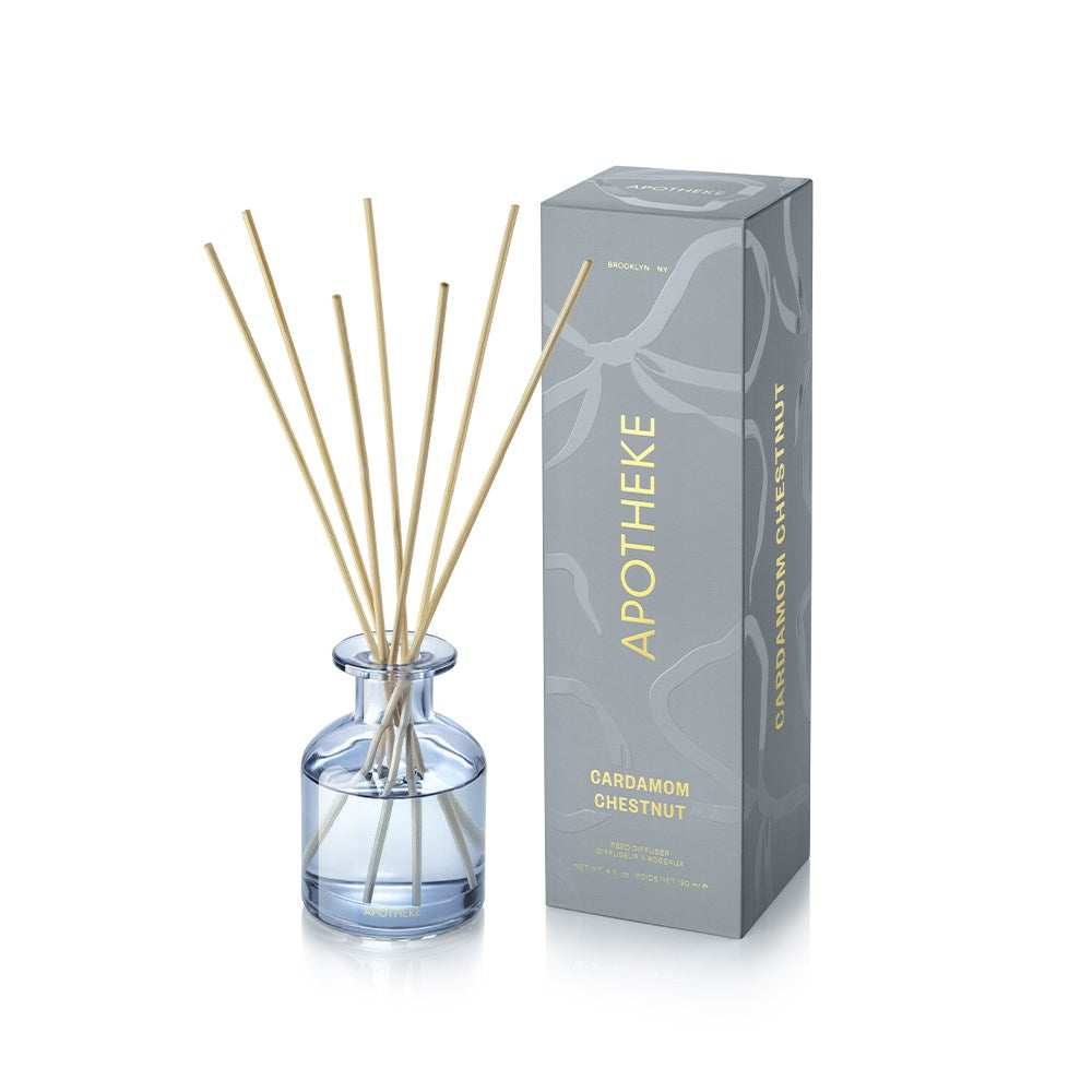 APOTHEKE Cardamom Chestnut Mini Reed Diffuser in a small blue-tinted glass bottle with pale liquid and fanned natural-colored reeds, beside a tall blue-gray patterned box with gold lettering.