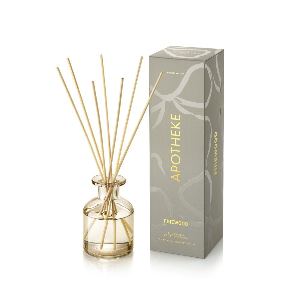APOTHEKE Firewood Mini Reed Diffuser in a small clear glass bottle holding pale golden liquid and fanned natural-colored reeds, beside a tall gray-green patterned box with gold lettering.