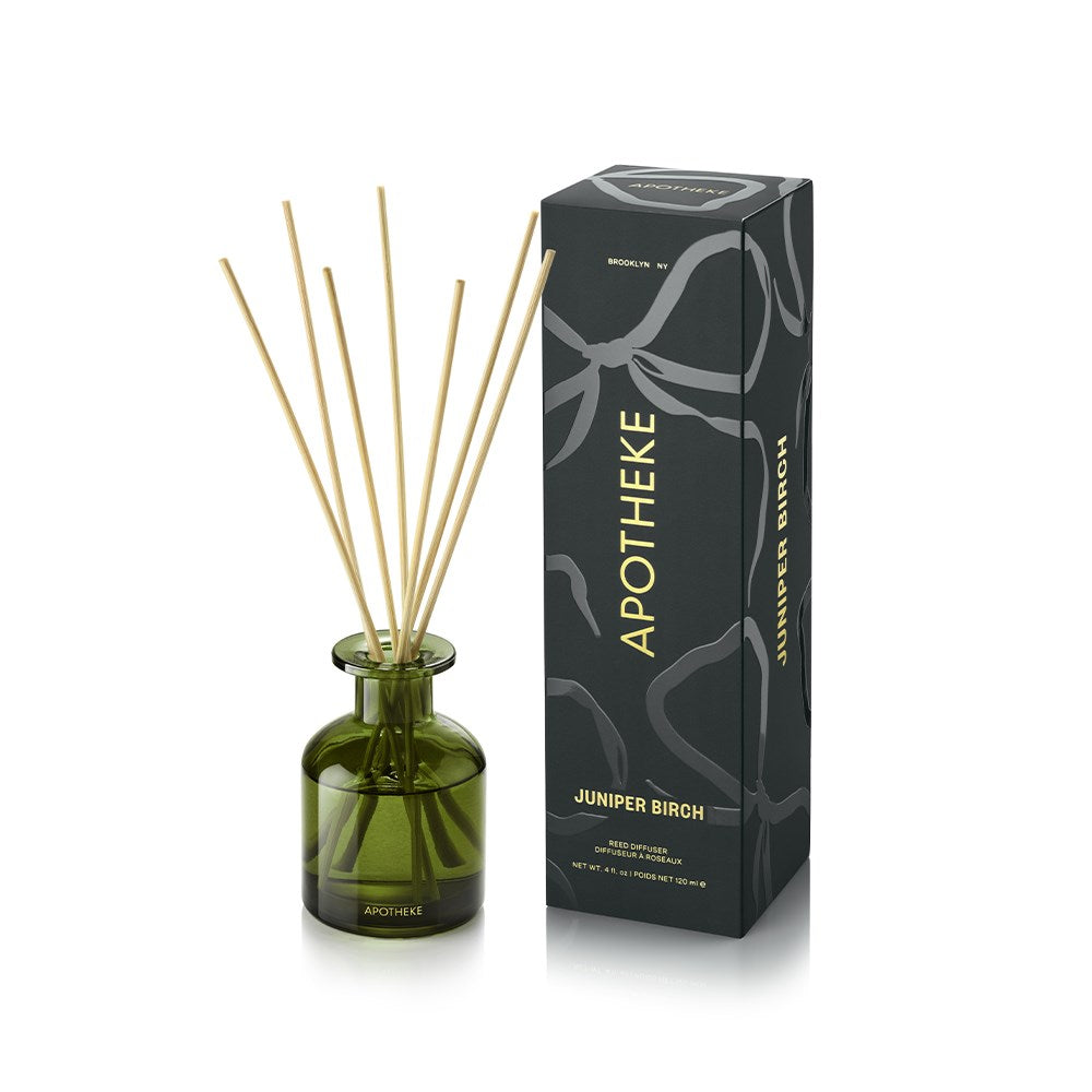 APOTHEKE Juniper Birch Mini Reed Diffuser in a green glass bottle with fanned natural-colored reeds, beside a tall dark green patterned box with gold lettering on a white background.