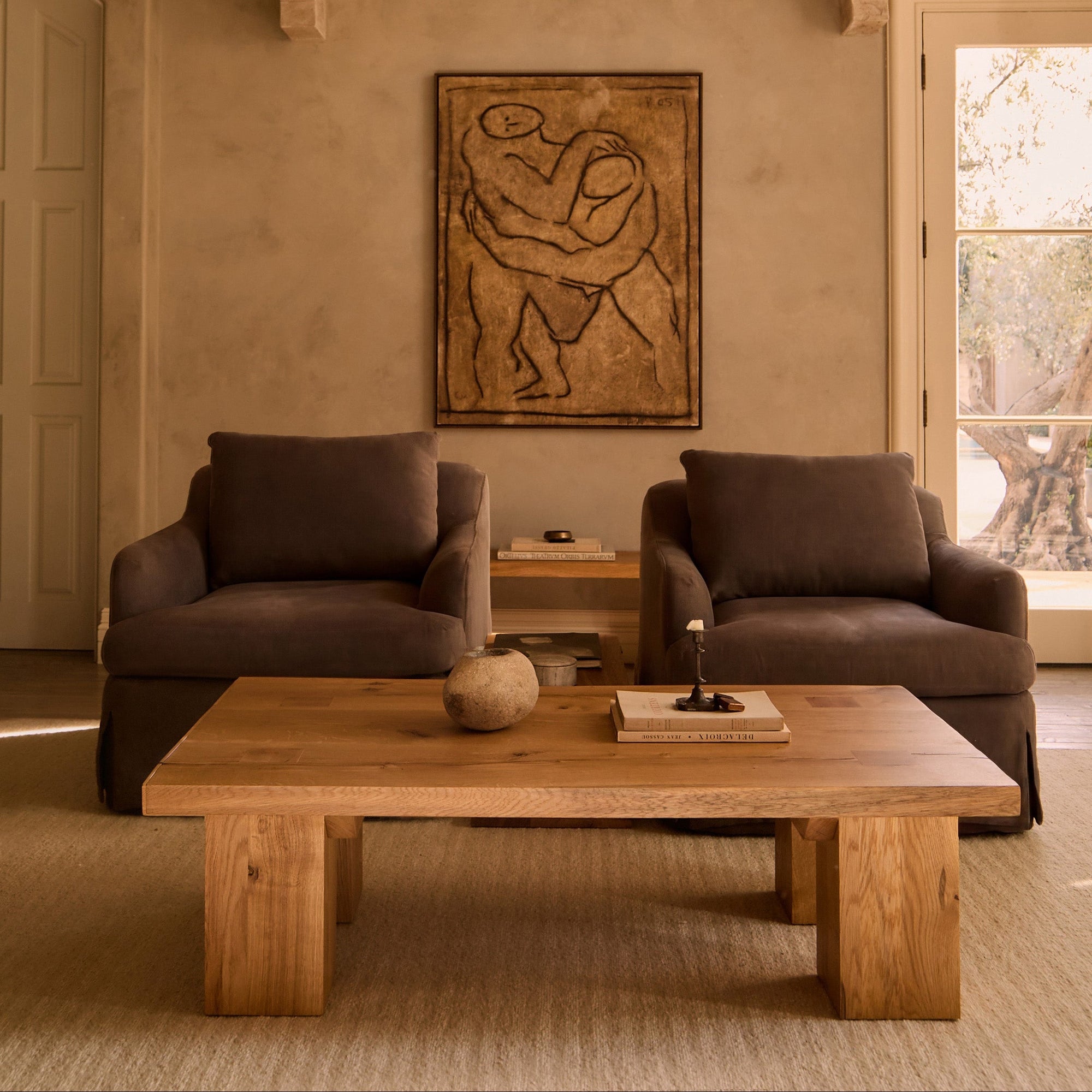 A warm, rustic living room scene featuring a large rectangular wooden coffee table in the foreground, flanked by two dark brown upholstered armchairs. A small wooden side table sits between them, and a large framed abstract painting hangs on the beige plaster wall behind by House of Leon.