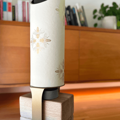 Close-up of the Apis tube and pale wood base in a warm interior, showing scattered metallic-gold bee motifs.