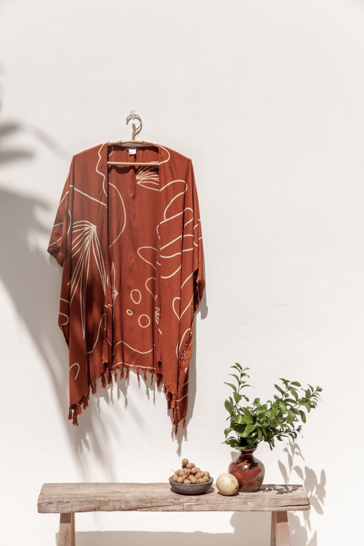 Village Thrive Desert Kimono, rust terracotta rayon, hanging from a wall hook above a wooden bench with a small potted plant.