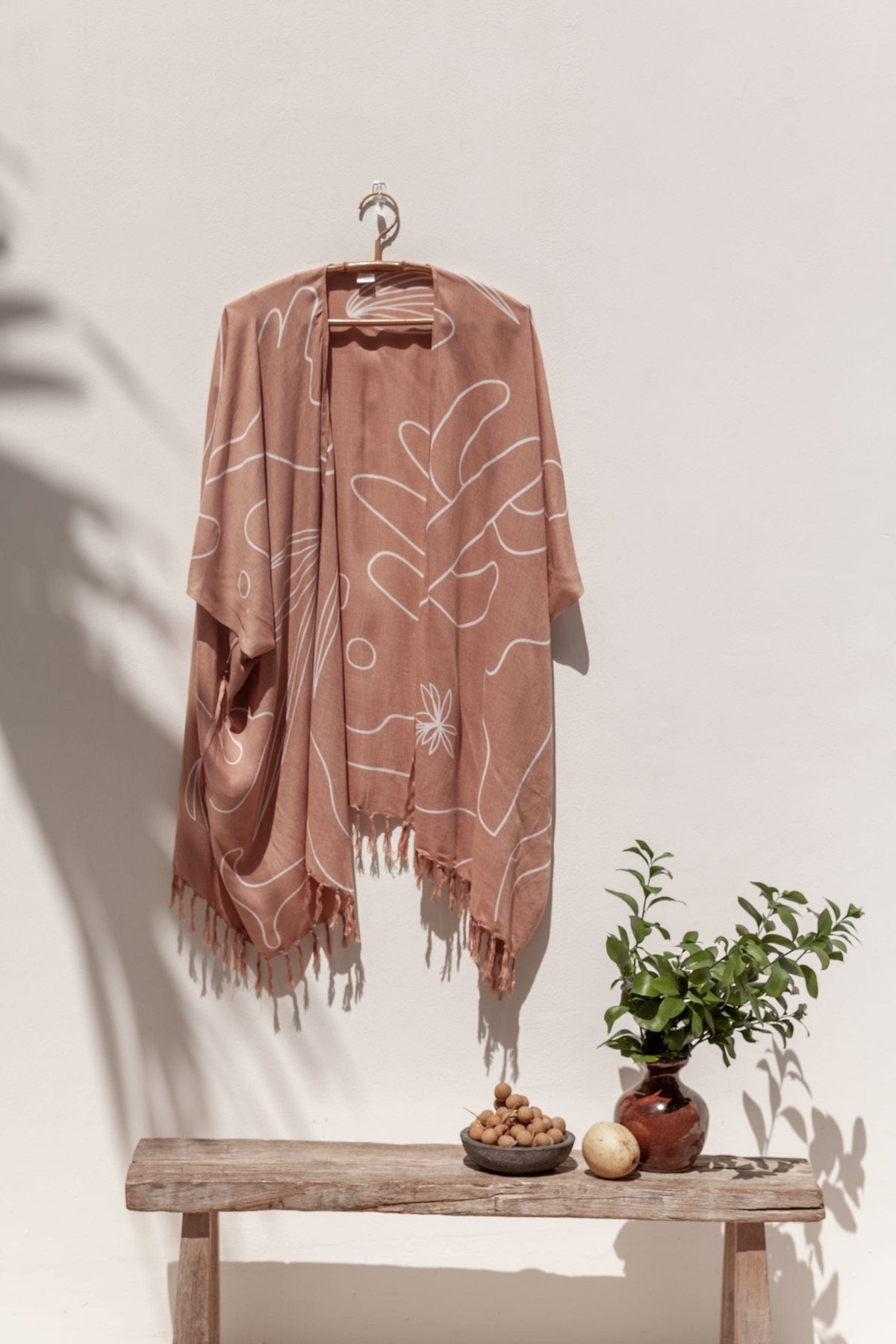 Village Thrive Desert Kimono, dusty rose rayon, hanging on a wooden hanger against a plain wall with a wood bench and small potted plant below.