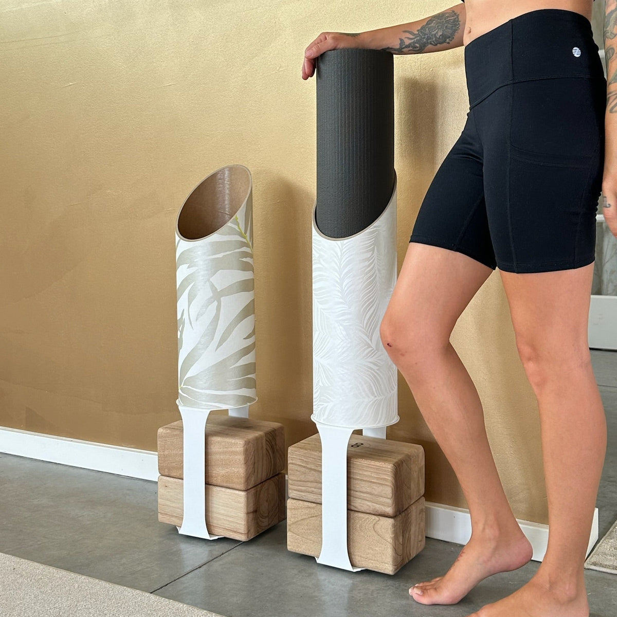 Woman in black activewear placing a rolled mat into a white patterned tube beside the plain white Sattva tube.