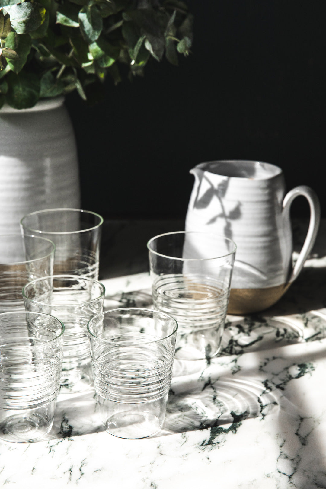 Group of clear ridged glasses beside a white ceramic pitcher on a dramatically sunlit marble table.