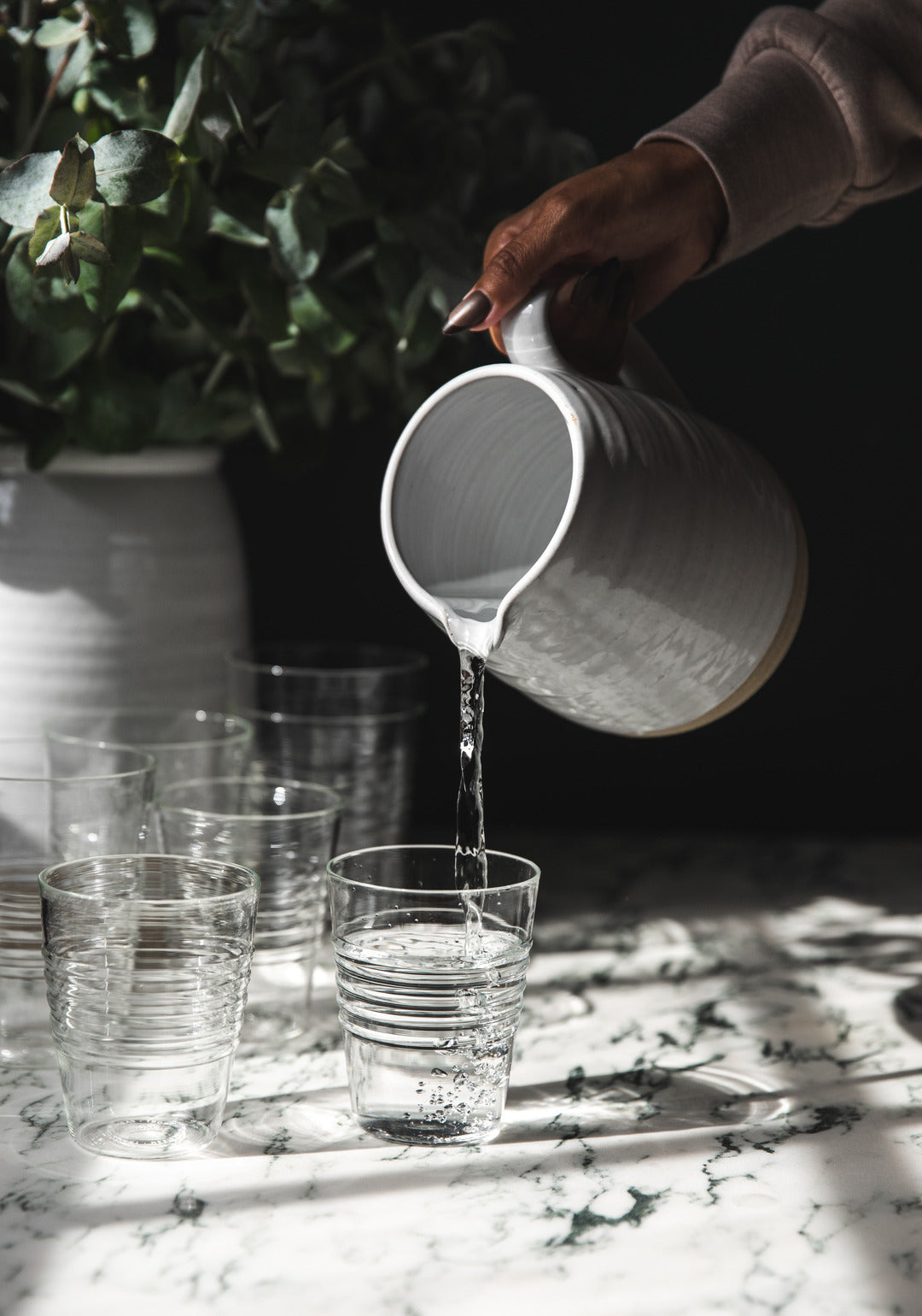Hand pouring water from a white ceramic pitcher into clear Farmer’s Glasses on a sunlit marble table.