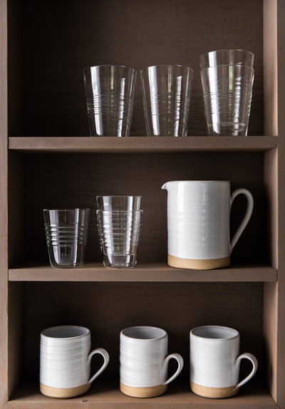 Clear Farmer’s Glasses displayed with white ceramic pitchers and mugs on dark wooden shelves.