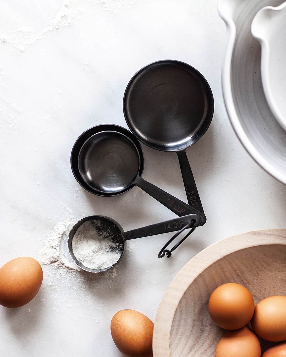 Black measuring cups holding flour beside brown eggs, a wooden bowl, and the curved edge of a white baking dish.