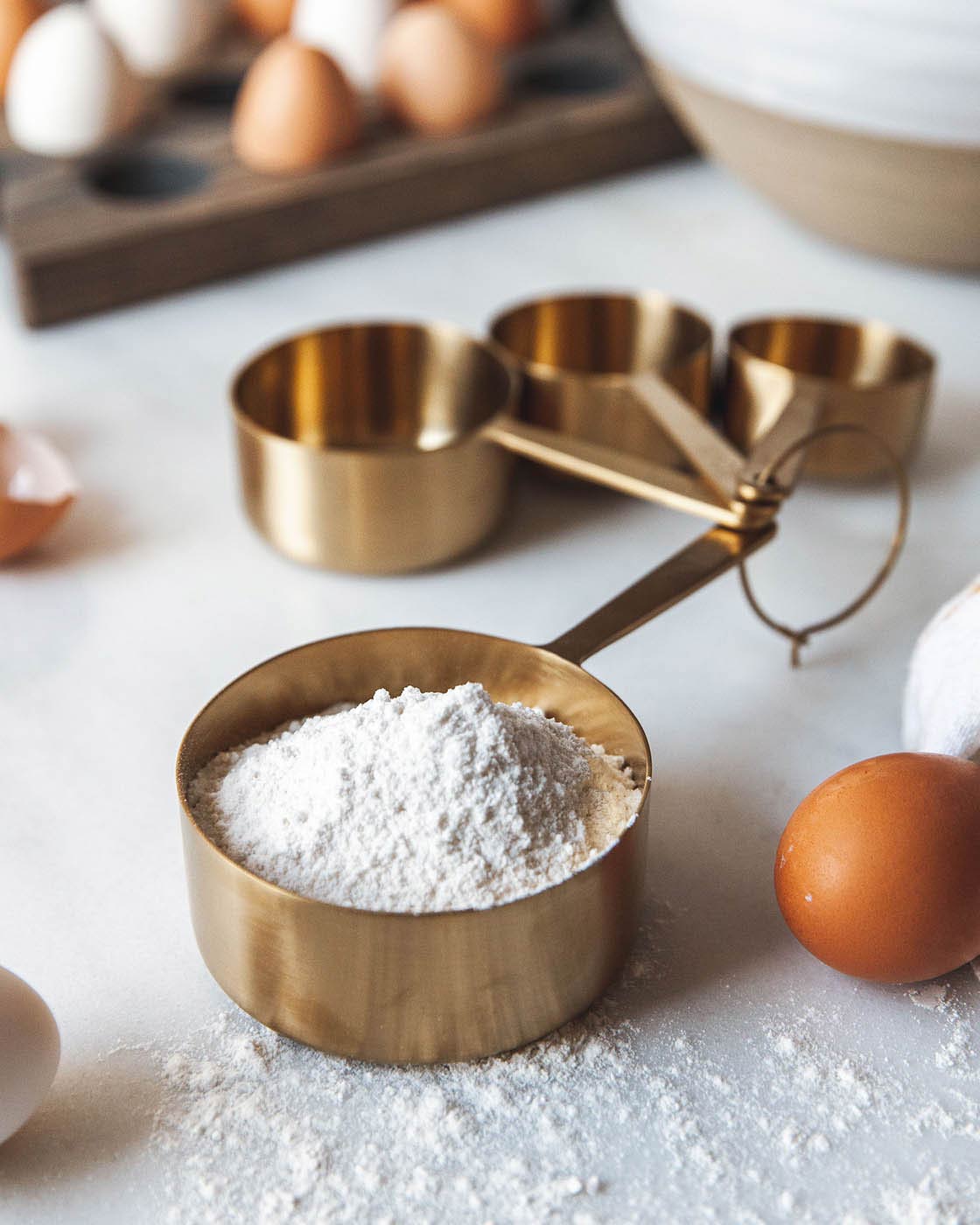Brass measuring cups used during baking, with the largest cup filled with flour beside brown eggs and a white mixing bowl.