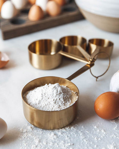 Brass measuring cups used during baking, with the largest cup filled with flour beside brown eggs and a white mixing bowl.