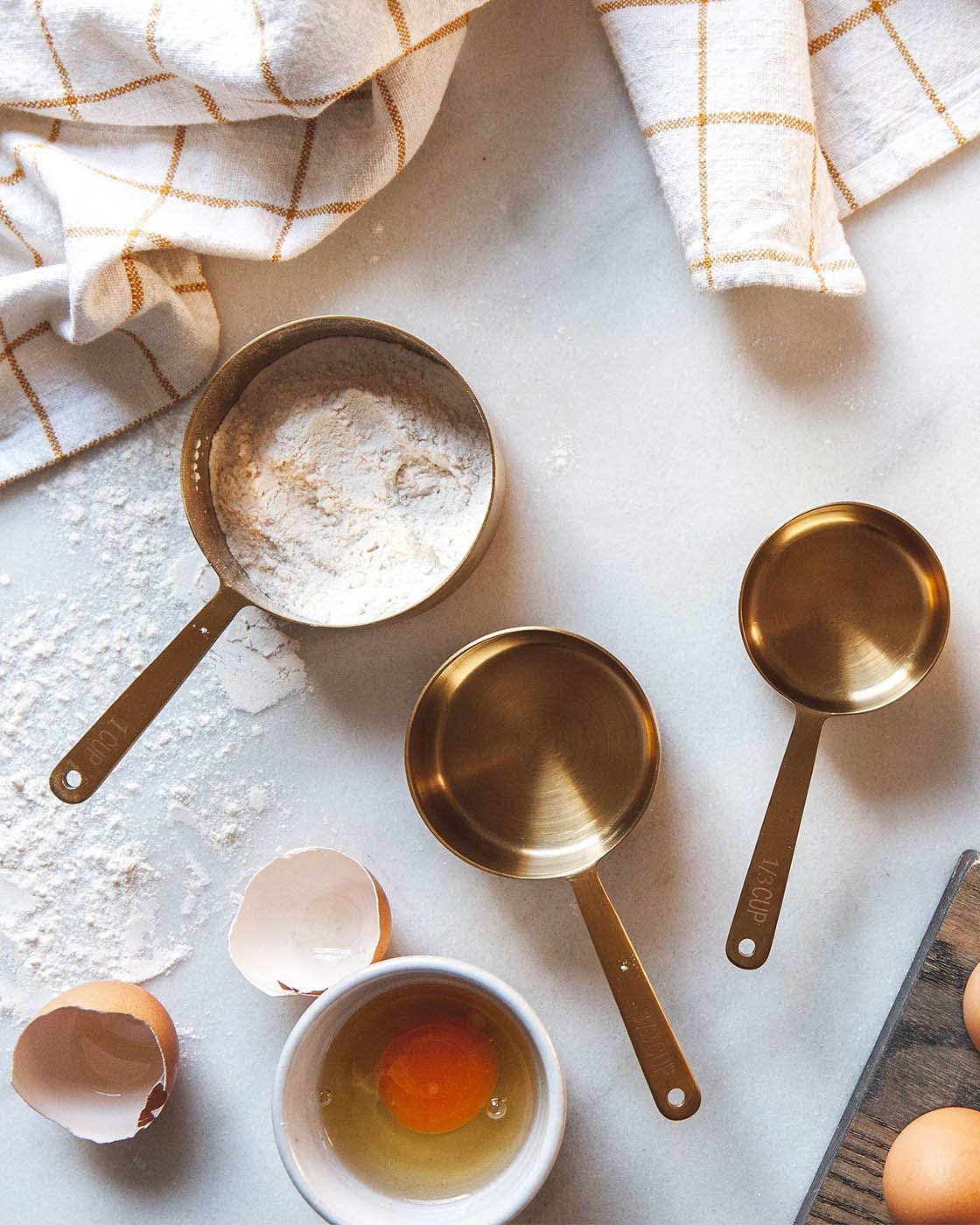 Brass measuring cups scattered across a flour-dusted work surface with cracked eggs, a whole egg, utensils, and folded striped towels.