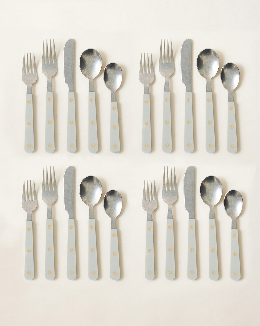Two complete pale blue-gray cutlery settings arranged side by side, each including two forks, a knife, and two spoons with triple-riveted handles.
