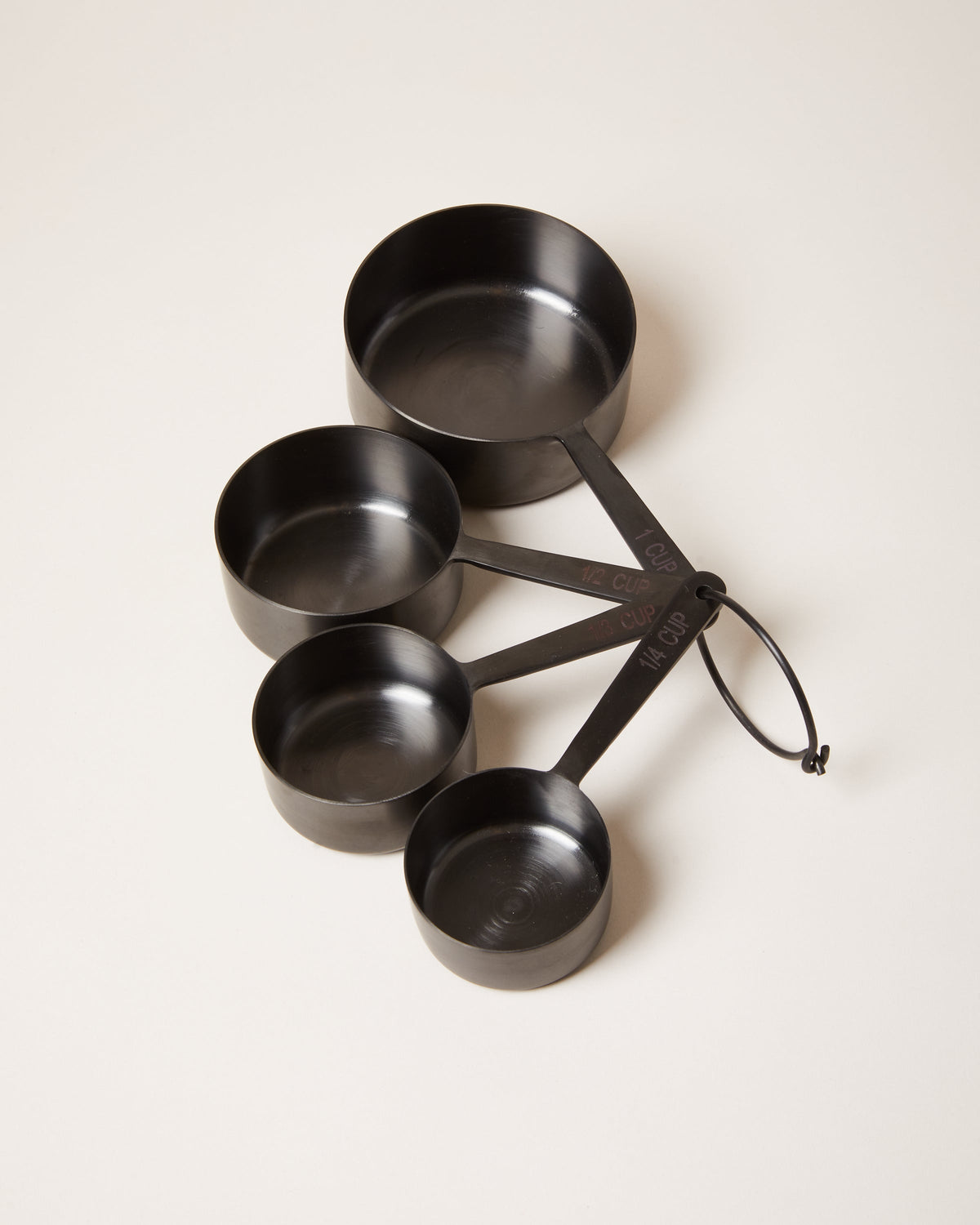 Four matte black measuring cups in graduated sizes, arranged in a fan and joined by a black storage ring.