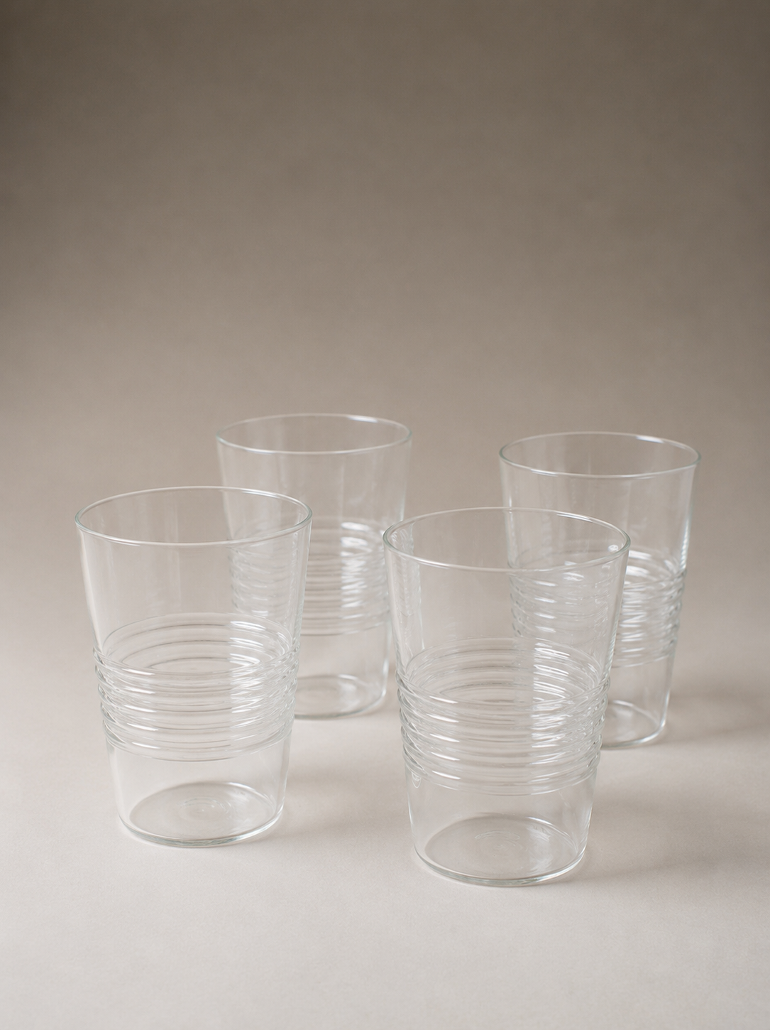 Four clear cylindrical drinking glasses with subtly irregular rims and horizontal ridges around their lower halves, arranged on a warm gray surface.