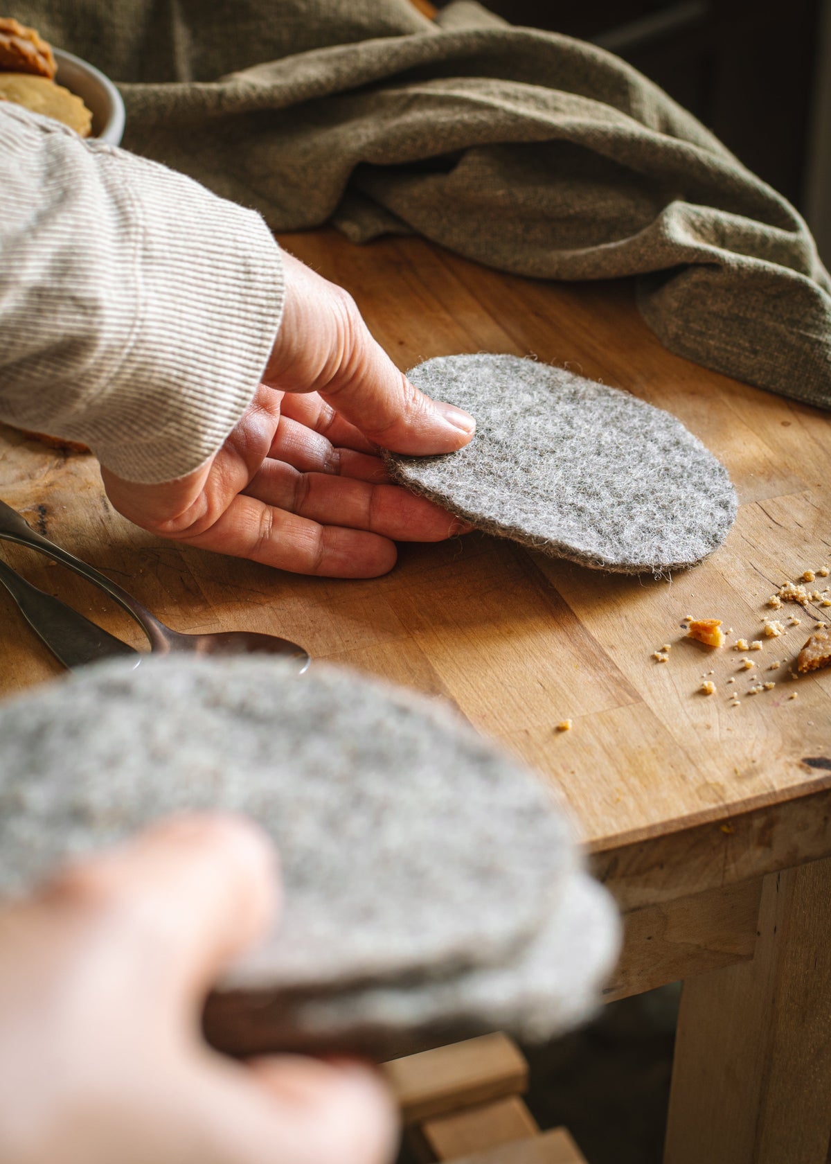 Hands placing a heather-gray felt coaster on a rustic wooden table while holding a small stack of matching coasters.