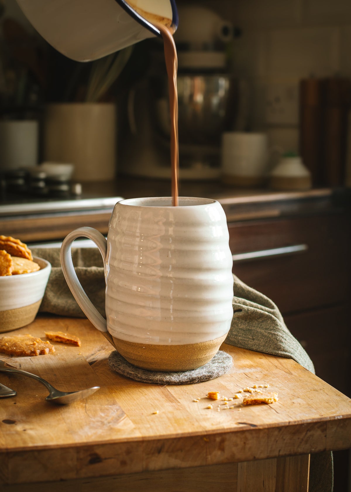 Dark coffee being poured into a white ribbed ceramic mug resting on a warm tan felt coaster atop a wooden counter.