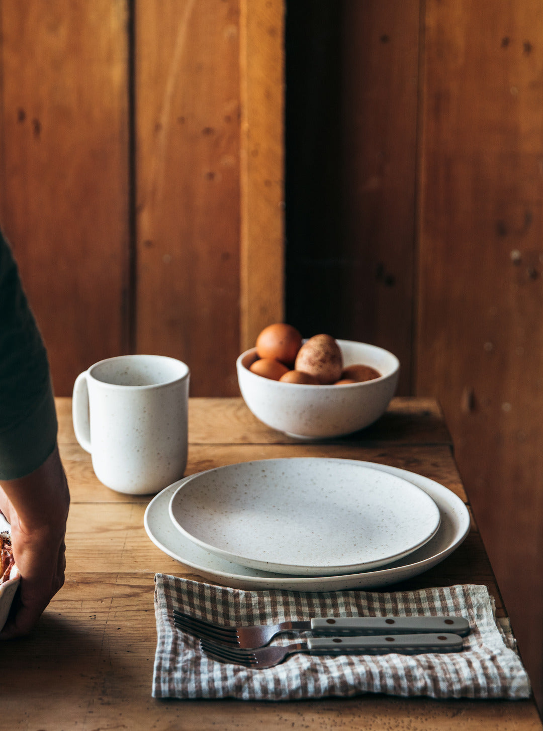 Blue-gray dinner plate and white mug arranged on folded checked linen beside a bowl of brown eggs on a rustic wooden table.