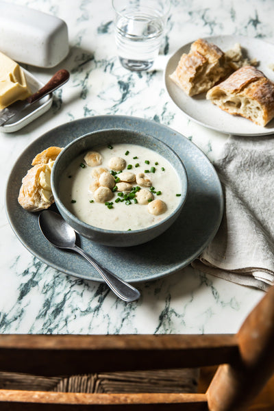 Blue-gray Fieldstone bowl filled with creamy soup, set on stacked plates with bread and silverware on a marble-topped table.