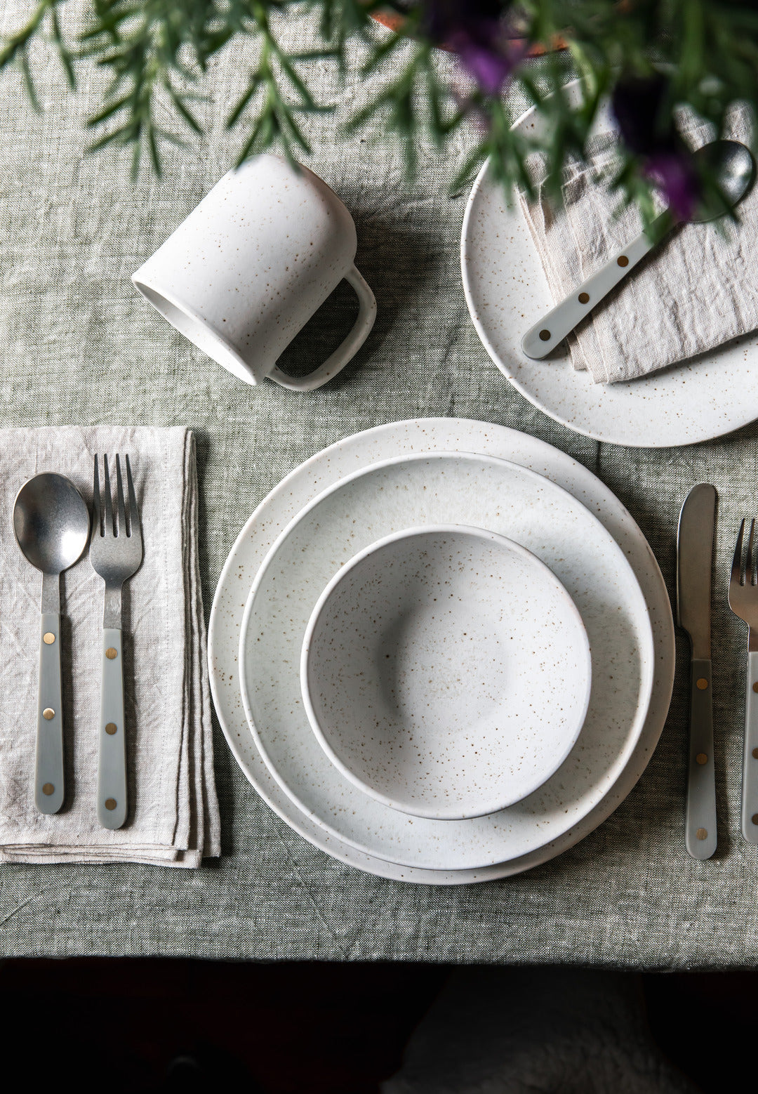 Overhead view of a white speckled bowl and stacked plates with a coordinating mug, silverware, folded gray napkins, and purple flowers.