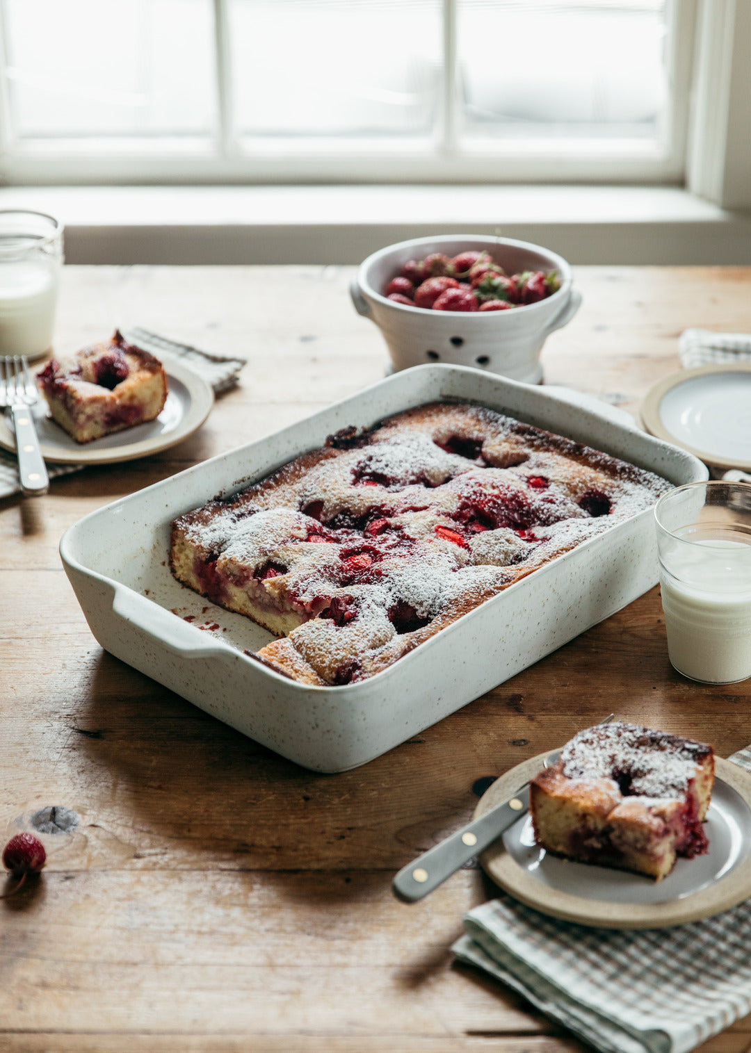 Berry cake in a white Fieldstone Baker on a rustic wooden table, surrounded by plated slices, red berries, folded napkins, and glasses of milk.