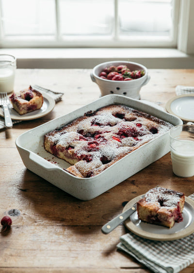 Berry cake in a white Fieldstone Baker on a rustic wooden table, surrounded by plated slices, red berries, folded napkins, and glasses of milk.