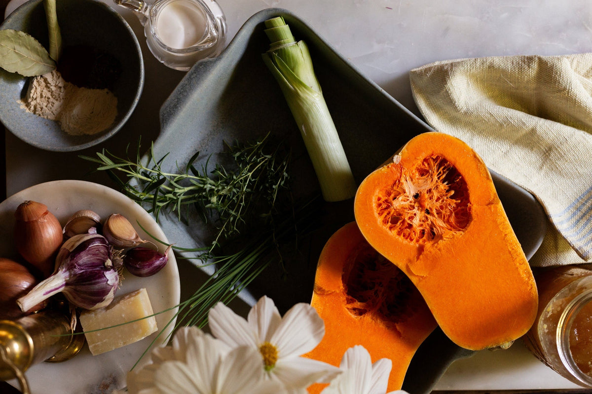 Overhead view of a blue-gray Fieldstone Baker filled with whole vegetables, including leeks and halved orange squash, surrounded by herbs, onions, and kitchen linens.
