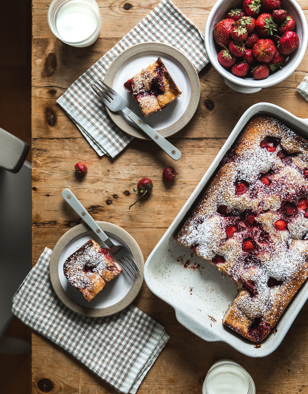 Overhead view of a berry dessert baked in a white Fieldstone Baker, dusted with powdered sugar and served beside bowls of berries and plated slices.