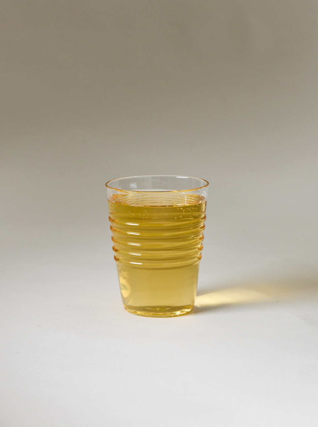 Small clear ridged glass filled with a translucent golden drink against a neutral studio background.