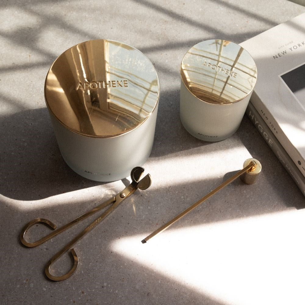 Gold wick trimmer and matching candle snuffer arranged beside two pale candle vessels with reflective gold lids on a gray surface crossed by sunlight and window shadows.