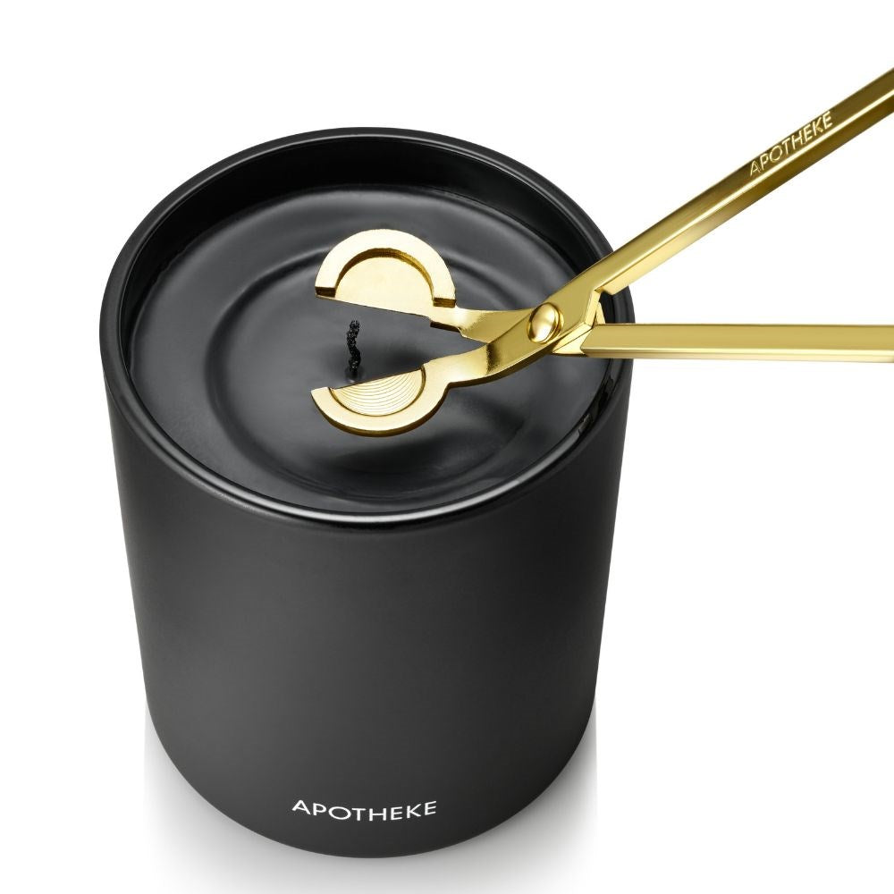 Gold wick trimmer positioned around the wick of an unlit black APOTHEKE candle, viewed from above with the cutting head resting just over the dark wax.