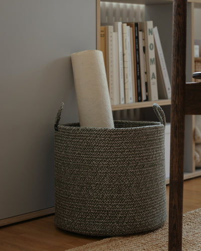 Tall rope basket filled with rolled materials beside a wooden bookcase.
