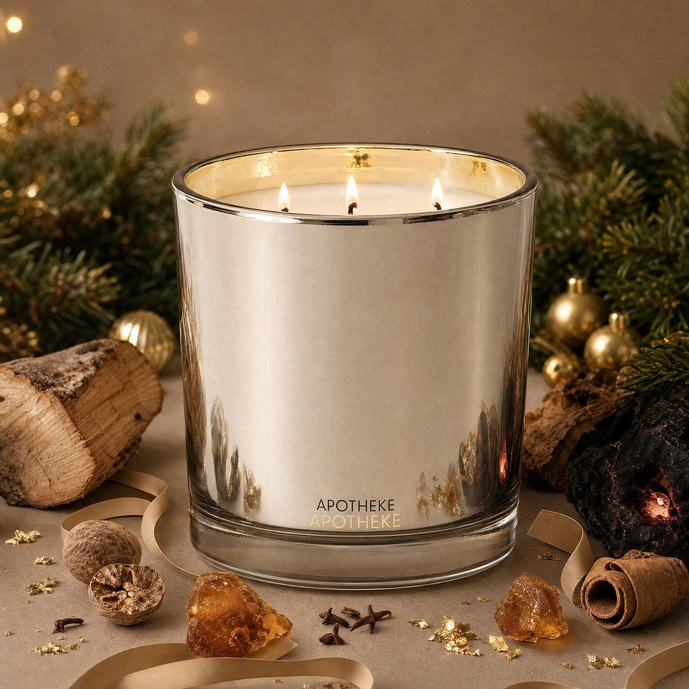 Three lit wicks glow inside the gold-toned Firewood candle, surrounded by evergreen branches, gold ornaments, wood pieces, spices, and amber-colored accents.